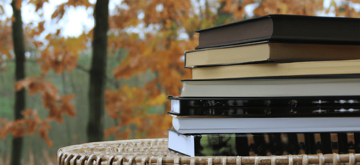 A photograph of a pile of fall books sitting on a wicker table with autumn trees in the background