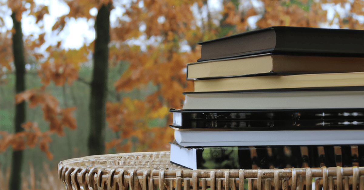 A photograph of a pile of fall books sitting on a wicker table with autumn trees in the background