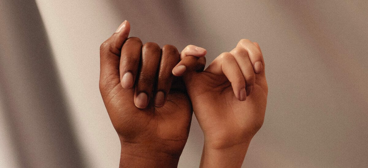 Two hands linking pinkies with a darker skinned hand on the left and a lighter skinned hand on the right in front of a coffee-colored background