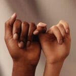 Two hands linking pinkies with a darker skinned hand on the left and a lighter skinned hand on the right in front of a coffee-colored background