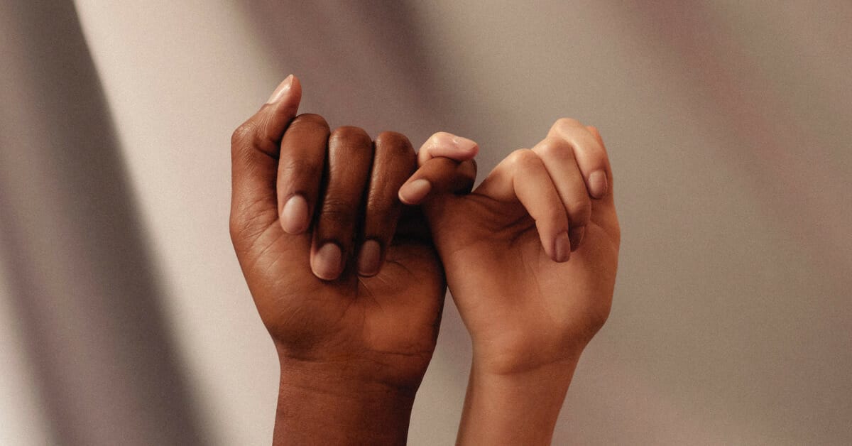 Two hands linking pinkies with a darker skinned hand on the left and a lighter skinned hand on the right in front of a coffee-colored background