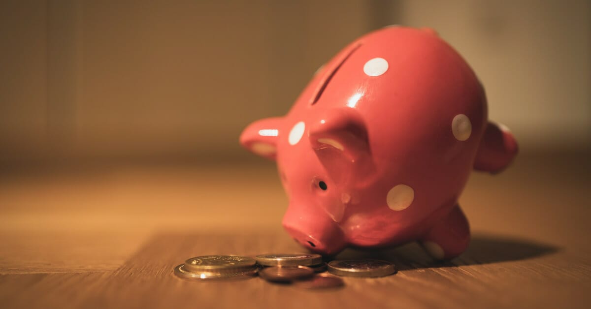 A pink ceramic piggy bank on a wooden table looking down nose-first at some scattered coins