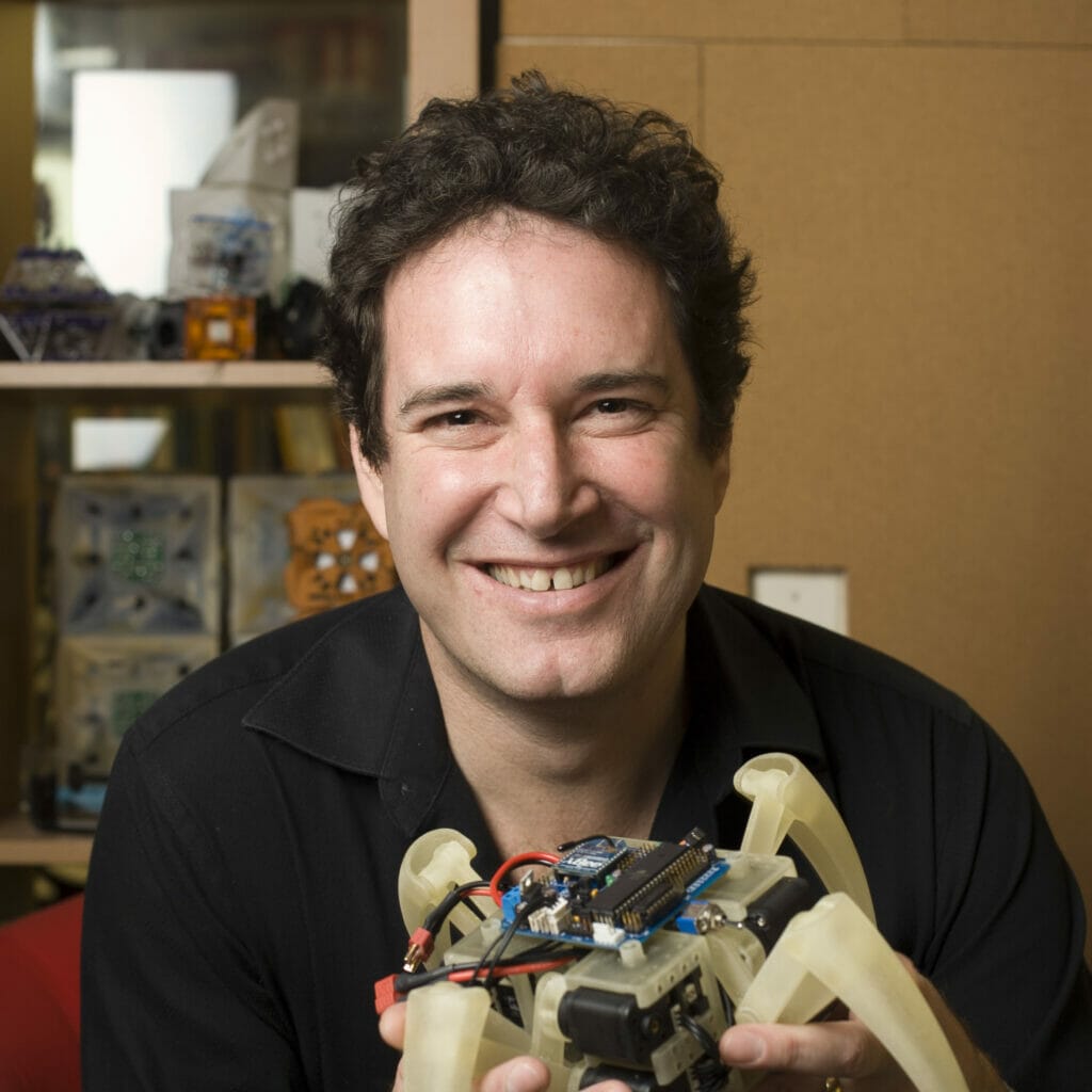 Hod Lipson smiling and holding a robot in front of a yellow wall