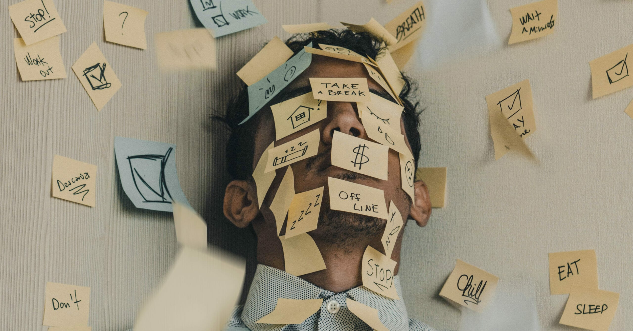 A man standing in a corner of two gray walls with multi-colored post-its all over his face. The post-its have tasks all over them.