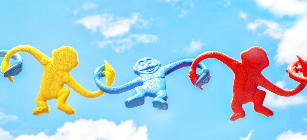 Yellow, blue and red plastic monkey toys have their arms linked together in front of a blue sky, symbolizing fun