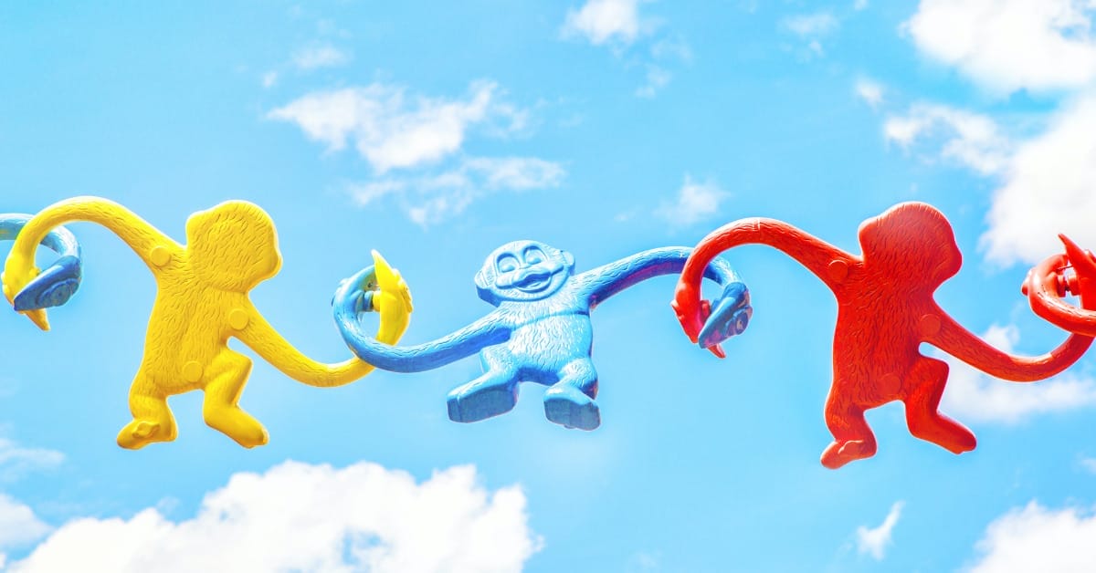 Yellow, blue and red plastic monkey toys have their arms linked together in front of a blue sky, symbolizing fun