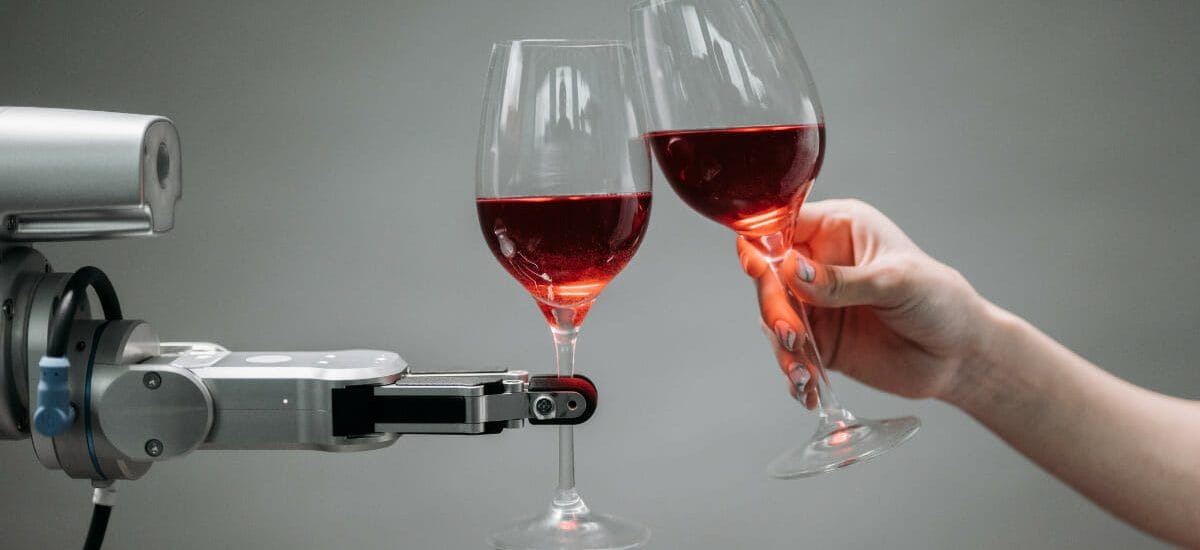 A robot hand holding a wineglass and clinking glasses with a human hand that is also holding a wineglass in front of a medium gray background