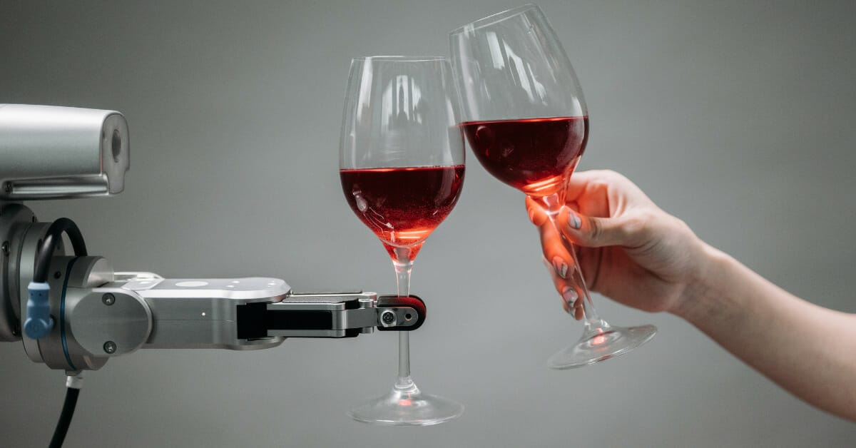 A robot hand holding a wineglass and clinking glasses with a human hand that is also holding a wineglass in front of a medium gray background