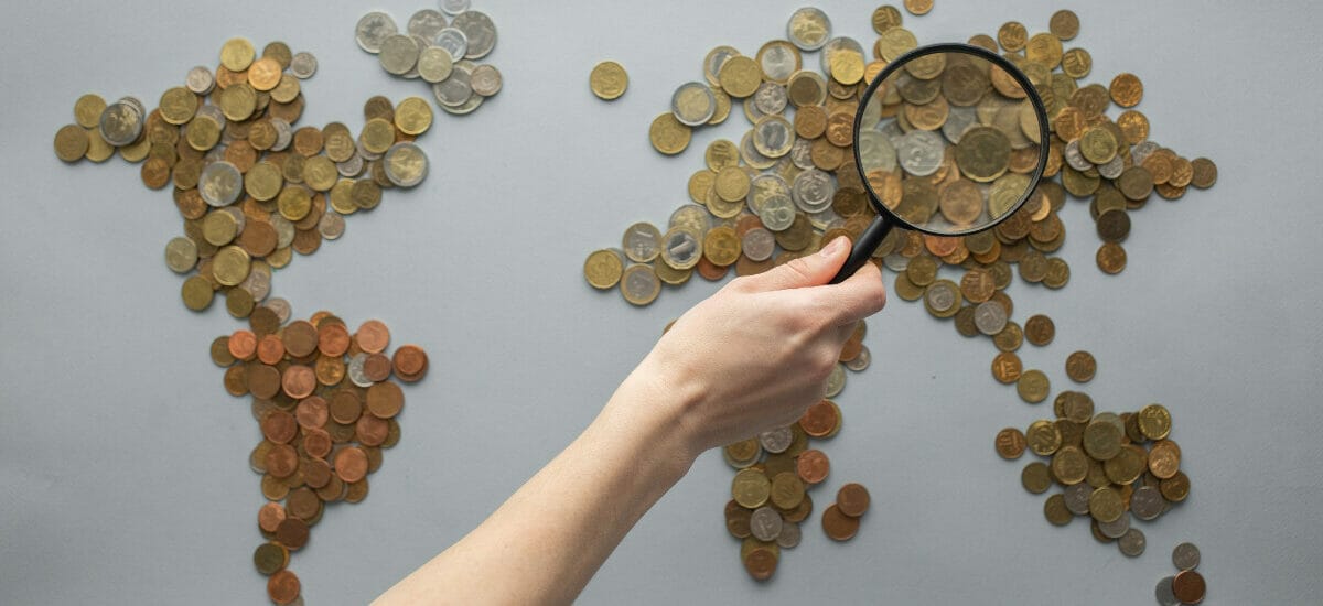 World map made of coins with a person’s hand holding a magnifying glass looking over the general region of Asia