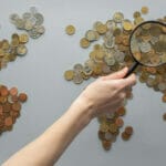 World map made of coins with a person’s hand holding a magnifying glass looking over the general region of Asia