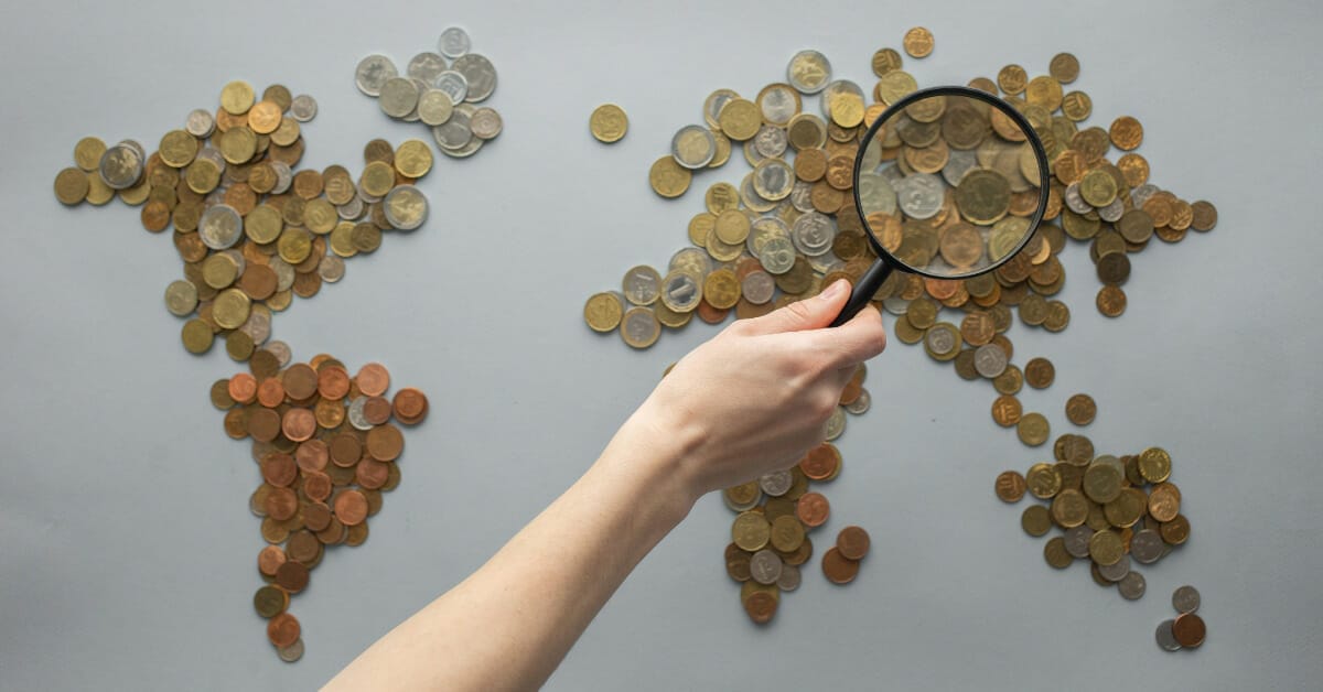 World map made of coins with a person’s hand holding a magnifying glass looking over the general region of Asia