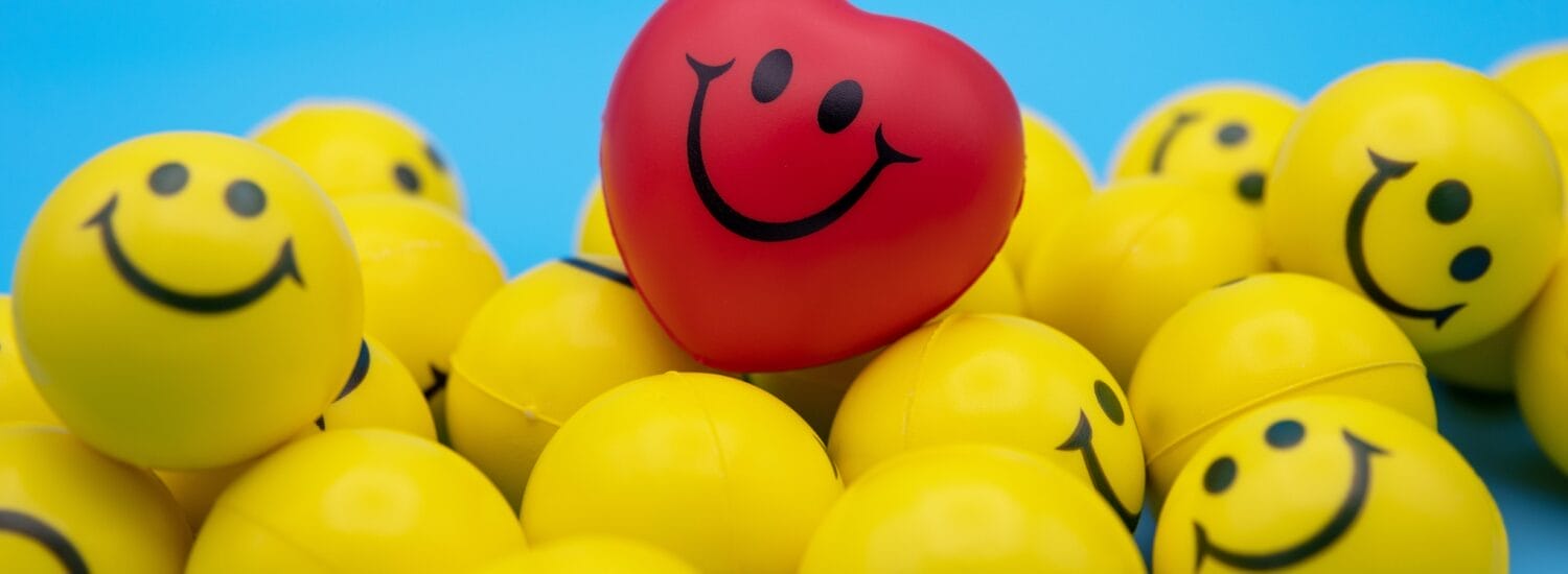 A group of yellow stress balls with happy faces on them in front of a bright blue background. There is one bright red ball with a happy face on top of the yellow ones.