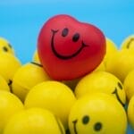 A group of yellow stress balls with happy faces on them in front of a bright blue background. There is one bright red ball with a happy face on top of the yellow ones.