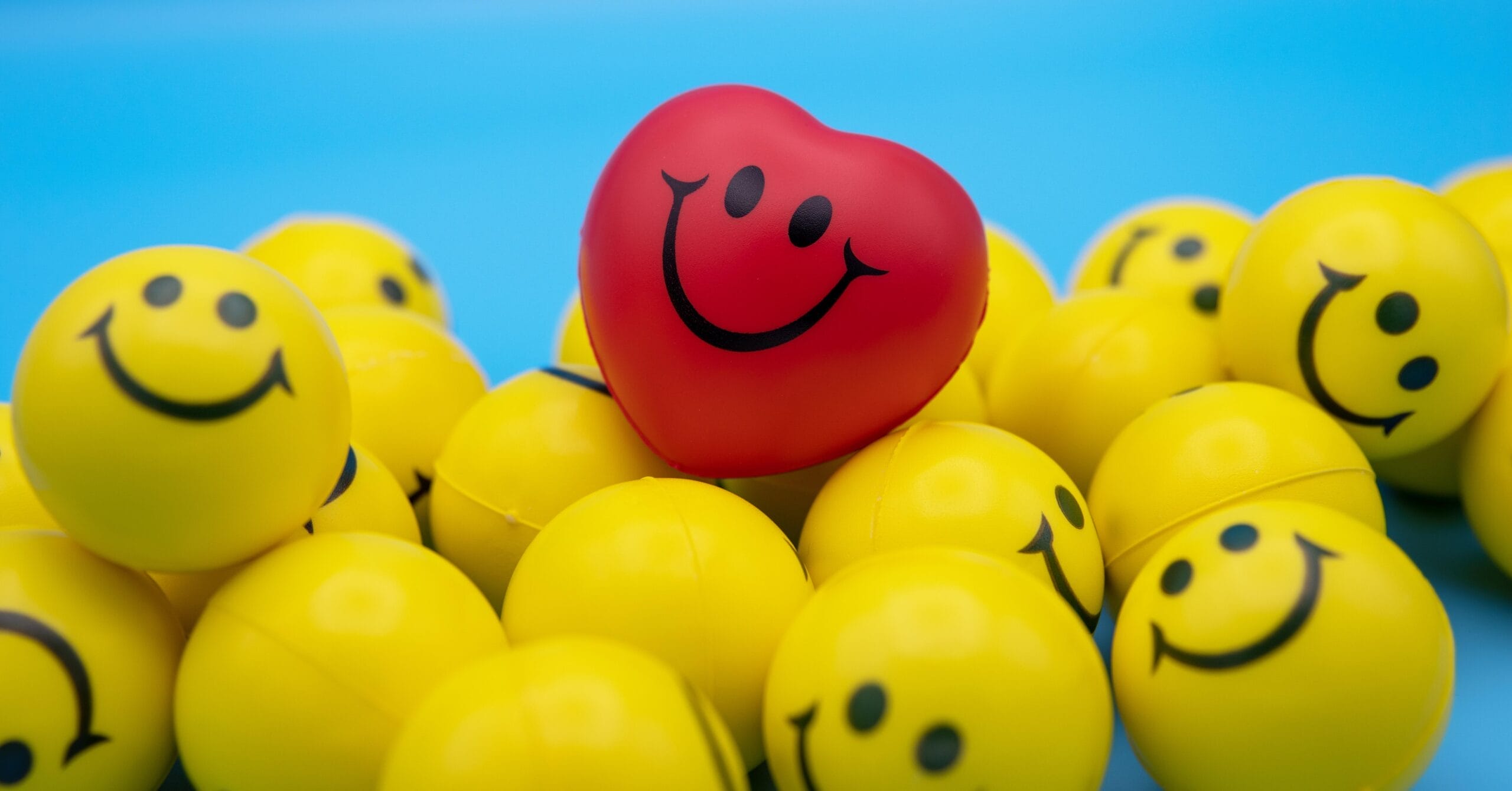 A group of yellow stress balls with happy faces on them in front of a bright blue background. There is one bright red ball with a happy face on top of the yellow ones.