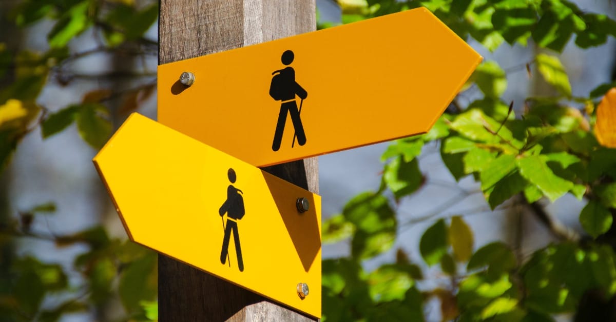 An orange arrow sign and a yellow arrow sign, both with black images of backpackers, pointing in two different directions, implying uncertainty, geopolitical or otherwise