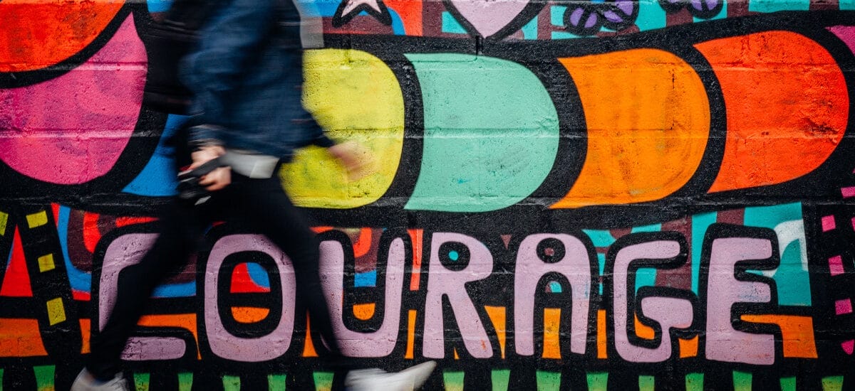 A man in blue pants and a dark blue jacket walking quickly in front of colorful graffiti that says "courage."