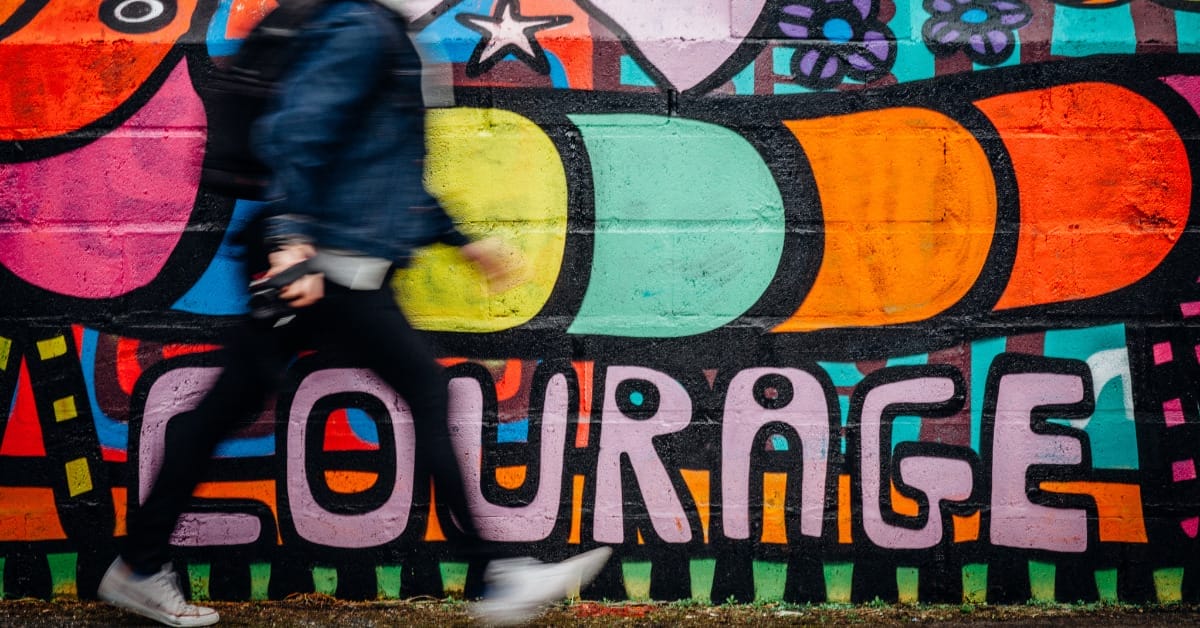 A man in blue pants and a dark blue jacket walking quickly in front of colorful graffiti that says "courage."