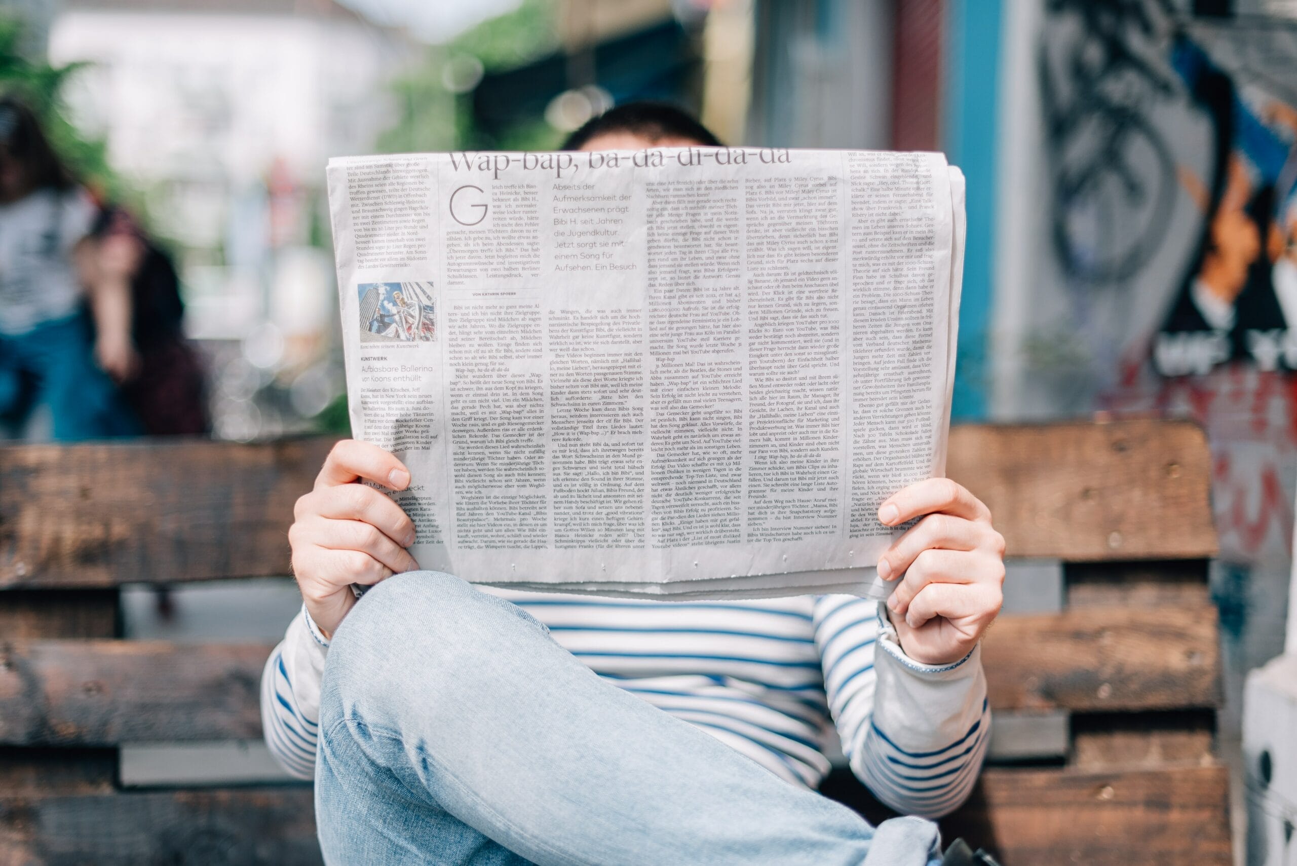 Person sitting on a bench holding a newspaper in front of their face