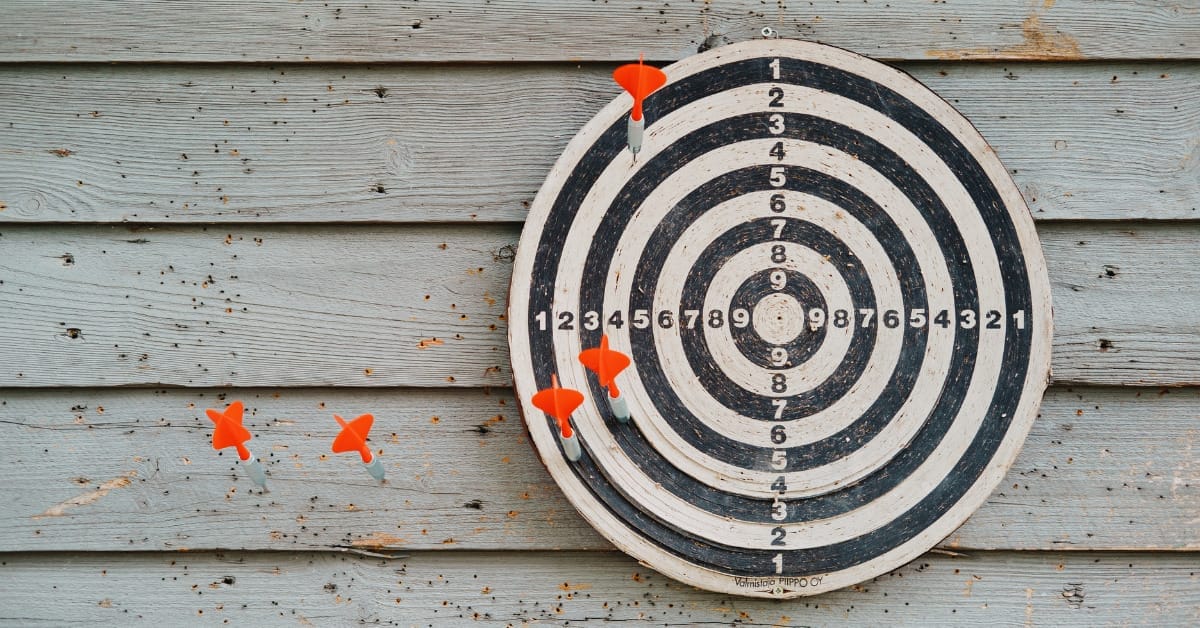 An image of a target with a bullseye target against a plain wooden wall. The dartboard has darts all aound it but none hit the target, implying failure