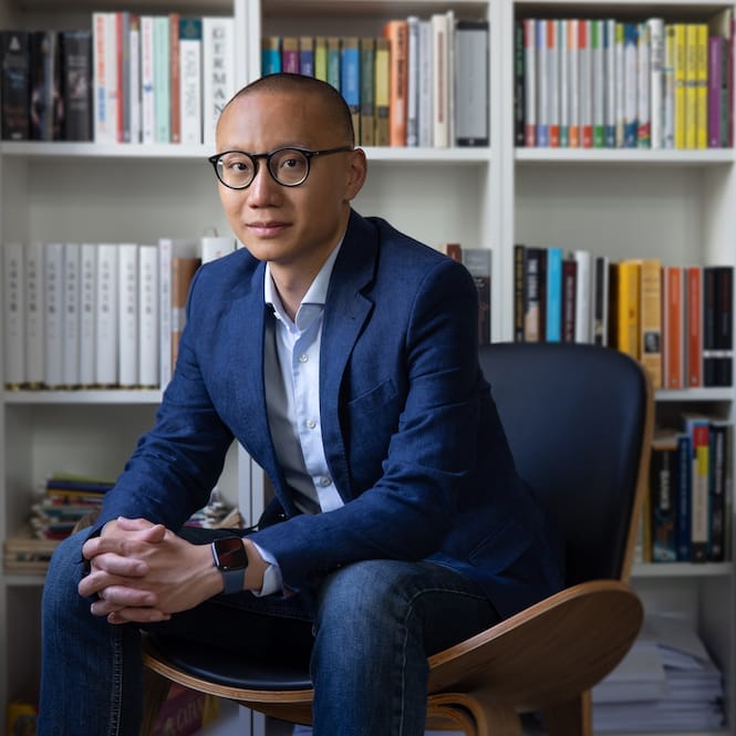 An image of Dan Wang in a blue suit sitting in a brown chair in front of a white bookshelf filled with colorful books. He has short hair, dark rimmed glasses and a lovely watch.